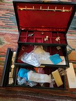 Full view of opened black jewelry box with red velvet interior showing various compartments filled with jewelry items including a gold-tone watch, small brooches, plastic bags with jewelry, and cleaning cloths.
