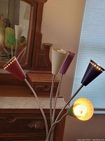 Photo of a multi-arm table lamp with five colored perforated lampshades placed on a wooden surface near a mirror and window blinds.
