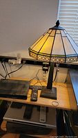 Tiffany style stained glass lamp with yellow and amber panels, sitting on a light wood table with electronics beneath and beside it