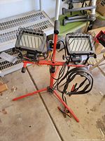 Dual head shoplight setup on a red tripod with attached power cord, showing lights with protective grills and sturdy construction.