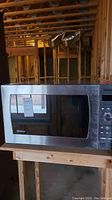 Closed Panasonic microwave with stainless steel front, digital control panel and dark glass door, sitting on wooden surface in unfinished basement.