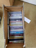 Box filled with multiple jewel case music CDs stacked in two rows showing titles on the spines