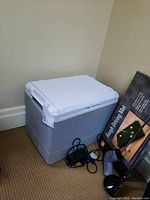 White and grey portable electric cooler seen from an angle showing the front and top sides with some electrical wires beside it.