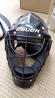 Front view of a black Bauer goalie helmet with a protective metal face cage and visible Bauer logo.