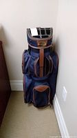 Front view of blue and brown golf bag standing upright, showing pockets and top club dividers.