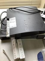 Sony PS3 console and Xbox 360 console stacked with two white Nintendo Wii controllers in front, showing cables and wear.