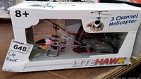 White and red LiteHawk 3 channel remote control helicopter in original box with cut-out window showing helicopter and transmitter. Box labeled for ages 8+. Front view taken on wood surface.