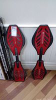 Photo showing two red and black Ripstik caster boards leaning against a wall on a wood floor, both boards showing wear.