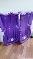 Two purple plastic sleds standing side by side on a wooden floor, showing wear and marks, with red pull ropes attached.