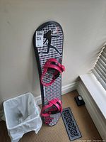 Front side view of the snowboard leaning against a wall next to a trash bin, showing the pink bindings and patterned design.