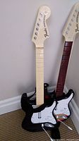 Front view of two black Rock Band video game guitars with cream colored necks, Fender styled headstocks, and white and sunburst pickguards.
