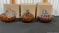 Three orange and black glass pumpkins displayed side by side on a black surface with brown cardboard boxes labeled "Dept Fall" in the background.
