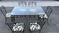 Full view of rectangular hammered glass tabletop with black wrought iron frame and six matching wrought iron chairs with floral cushions outdoors on pavement.