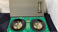 Two black lacquerware plates with hand-painted hummingbird and floral designs, plus two gold tone forks on a green satin-lined presentation box.