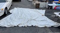 Full view of the large white canvas boat cover spread out on asphalt showing size and general condition with some wrinkles and dirt.