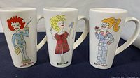 Front view of the three mugs showing cartoon woman designs, arranged side by side on a dark surface with white background.
