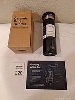Photo of the ceramic burr grinder box with label and the black French Press coffee to go cup with a usage instruction card on white surface against beige wall.