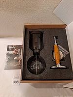 Top-down view of the boxed siphon coffee maker kit showing glass part and wooden handle stand tightly fitted in foam padding.