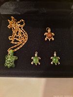 Gold tone chain necklace with green stone turtle pendant, one gold tone brown stone turtle charm, and two silver tone green stone turtle charms displayed in black case.