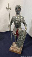 Front view of vintage miniature metal suit of armor with spear and shield, placed on wooden base.