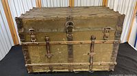 Full front view of vintage travel trunk showing metal reinforcements, two straps, latches, and central lock.