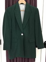 Front view of green blazer jacket hanging on wooden hanger, showing notch lapel, one white button, flap pockets and smooth fabric finish.