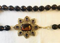 Close-up of the decorative clasp with topaz rectangular center stone and purple teardrop surrounding stones, with knotted smoky topaz crystal beads on either side.
