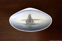Clam shell shaped trinket dish showing sailboat painting in blue and gray on white porcelain surface.