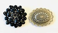 Two vintage brooches shown together on white background, one black faceted cluster and one silver embossed oval.