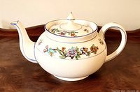 Side view of the vintage Aynsley teapot showing the white china body, blue trim along the edges, handle, and spout, and colorful floral band wrapping the body.