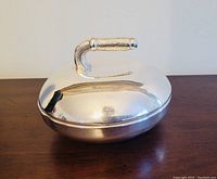 Photo of vintage silver-plated curling stone-shaped serving dish with lid and rounded handle, showing front and top views. Reflective polished surface with slight wear visible.
