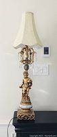 Full view of the finished lamp showing the cream fabric shade with subtle discoloration, hanging crystal pendants, and detailed gold cherub figure on a decorative base.