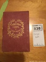 Front cover of the antique Christmas Carol book by Charles Dickens, showing red cloth with gold embossed text and wreath design.