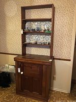 Full view of the two-piece wooden sideboard showing lower cabinet with door and drawer, and open shelving unit on top with decorative plates on shelves (not included). Cabinet placed against wallpapered wall on wood patterned floor.