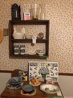 Full view of wooden wall shelf with ceramic, glass, and metal items, and table below with dishes and placemats.