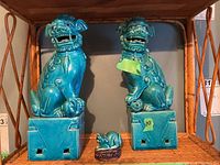 Front view of two large turquoise ceramic Foo Dog figurines sitting on square pedestals with a small figurine in front. One large dog has label '10'.