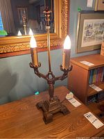 Full view of metal lamp base with two candle-shaped bulbs lit, showing ornate base detail and full height with the wicker shade attached.