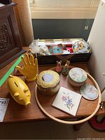 Photo of lot items on small round wooden table including yellow ceramic piggy bank, yellow ceramic hand figurine, two rabbit figurines, decorative tins, floral tile coaster, and sewing notions tin.