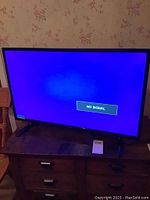 RCA TV displaying 'No Signal' on screen while turned on, placed on wooden stand with floral wallpaper background.