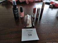 Seven cosmetic items on table with MaxSold lot number card - setting spray, nail polish, lipsticks, mascara, concealer, foundation stick