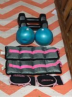 Top down view of exercise equipment set including dumbbells, two blue Bally Fitness balls, pink-accented ankle weights, and Piloxing gloves on patterned rug
