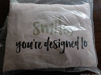 White pillow with 'Smile, you're designed to' text in black and glittery gold print, packaged in clear plastic.