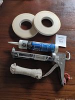 Two rolls of double sided tape, a tube of GE Power 8 Silicone Caulking, a metal caulking gun tool, and a coiled white rope on wooden surface.