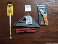 Photo showing DeWalt hacksaw blade package, black plastic speed square, orange Stanley utility knife in package, two red carpenter pencils on wooden surface.