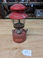 Front view showing the red metal body, glass globe, control knob, and Coleman badge on base with rust and wear.