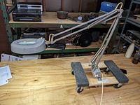 Full view of the Luxo articulated lamp on a wooden table with shelves in the background, showing the lamp's arm and base with the mounting bracket and caster trolleys.