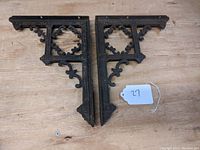 Pair of black cast iron Eastlake shelf brackets placed side by side on wooden surface with a white tag labeled '27'.