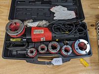 Overview of the entire lot showing the red and black portable electric threading machine labeled X1003, six threading dies, carrying case, gloves, and other threading accessories.