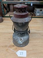 Front view of a heavily rusted vintage lantern with clear glass globe and red metal top, showing the knob and base details