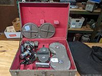 Open carrying case showing camera body with attached lens, three film magazines, power cable and keys on red felt interior